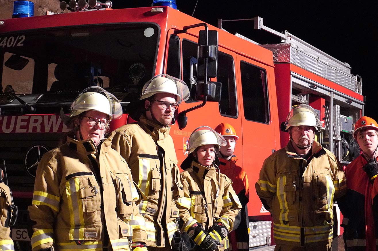 Mehrere Personen und beiger Feuerwehruniform mit Helmen stehen vor einem roten Feuerwehrfahrzeug.