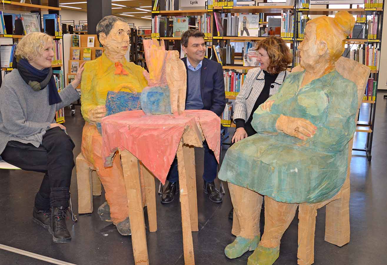 Holzskulptur mit zwei Damen, um die herum drei Personen sitzen