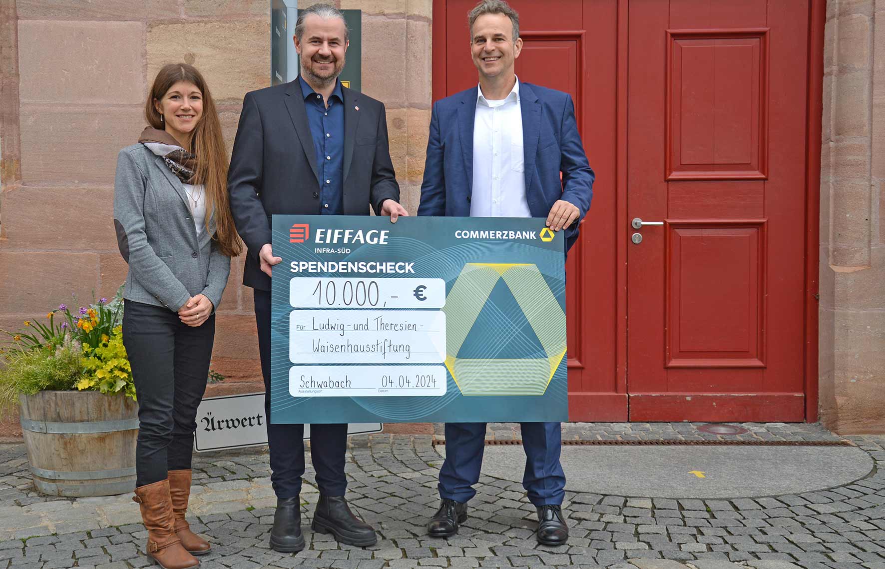 Eine Frau und zwei Männer stehen vor der dunkelroten Rathaustür. Sie halten einen überdimensionalen symbolischen Scheck über 10.000 Euro in Händen.