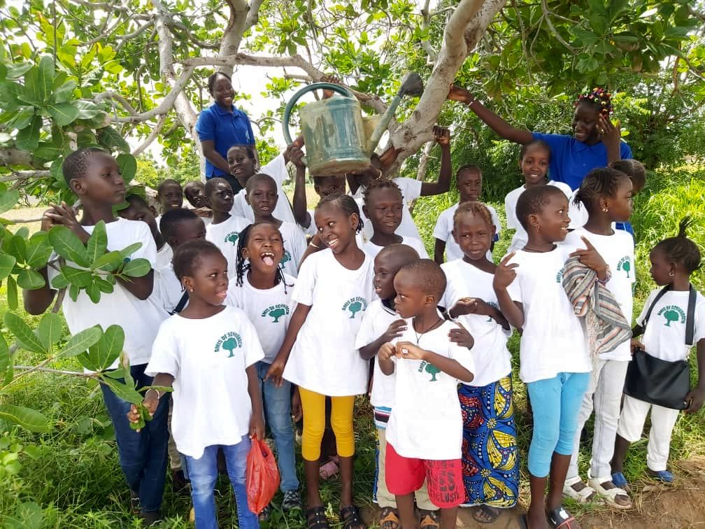 Kinder aus Gossas stehen unter einem Baum und halten Gießkannen und andere Gartenwerkzeuge in den Händen.