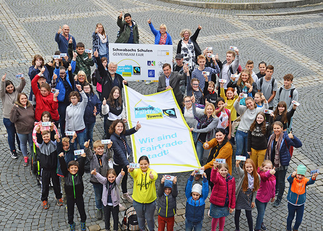 Alle Beteiligten halten eine Fairtrade-Fahne hoch und freuen sich.