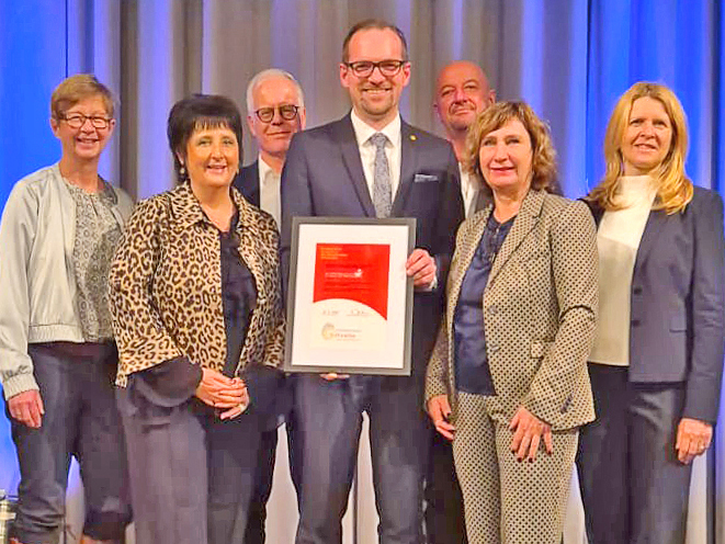 Eine Gruppe von sieben Personen steht vor einem blauen Vorhang. Die Personen tragen formelle Kleidung, darunter Anzüge, Blazer und gemusterte Jacken. Die Person in der Mitte hält einen gerahmten roten und weißen Urkundenrahmen mit beiden Händen vor der Brust. Die Personen stehen eng beieinander und blicken in Richtung Kamera.