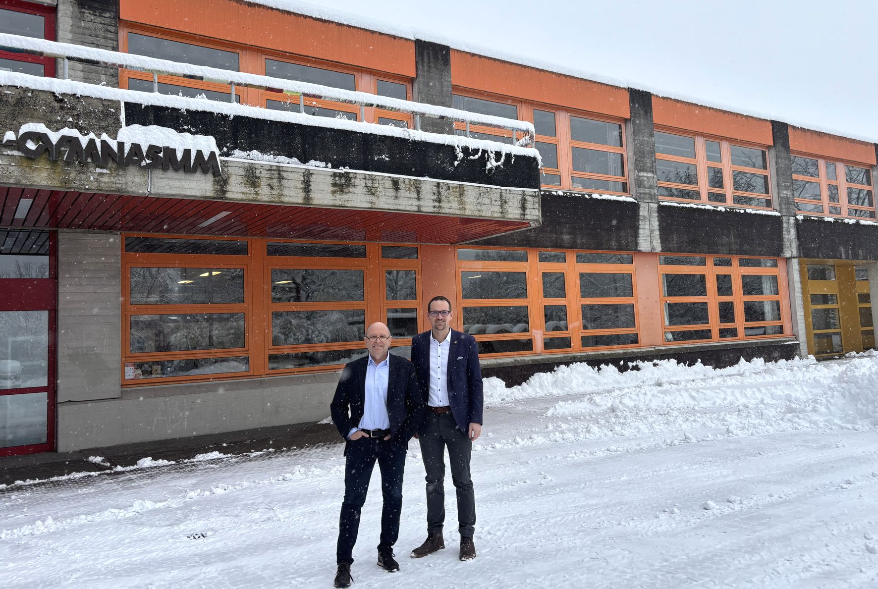 Zwei Männer in Anzügen stehen vor einem orangefarbenen Schulgebäude mit Schnee auf dem Boden und dem Dach