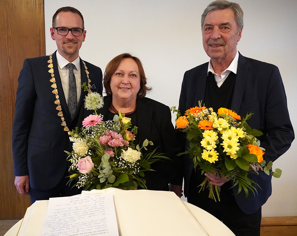 Drei Personen stehen nebeneinander, zwei halten große Blumensträuße, eine trägt eine Amtskette, vor ihnen liegt ein aufgeschlagenes Buch mit handgeschriebenem Text