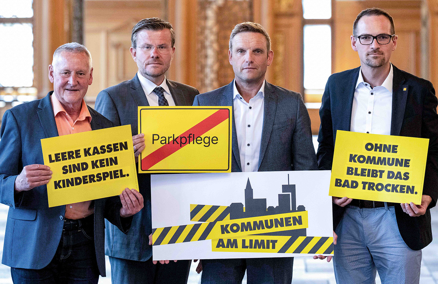 Vier Männer in Anzügen halten gelbe Schilder mit Texten zu kommunalen Problemen, darunter 'LEERE KASSEN SIND KEIN KINDERSPIEL', 'Parkpflege' durchgestrichen, 'OHNE KOMMUNE BLEIBT DAS BAD TROCKEN' und 'KOMMUNEN AM LIMIT' vor einer Holzvertäfelung.
