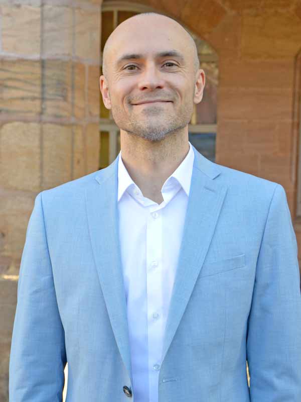 Mann in blauem Anzug steht vor einem Sandsteingebäude. 