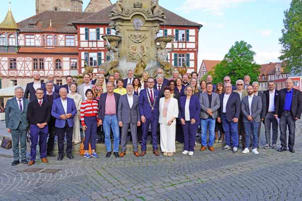 43 Männer und Frauen stehen in Reihen vor einem Brunnen. Dahinter sieht man ein Fachwerkhaus mit einem vergoldeten Erkerturm.