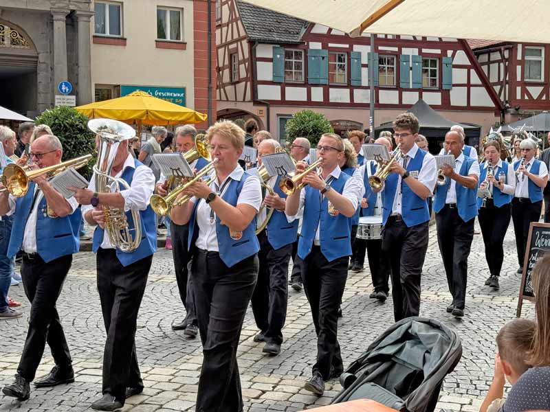 Musiker und Musikerinnen mit Trompeten, Posaunen, Tuben und anderen Blasinstrumenten und blauen Westen ziehen musizierend über den Marktplatz.