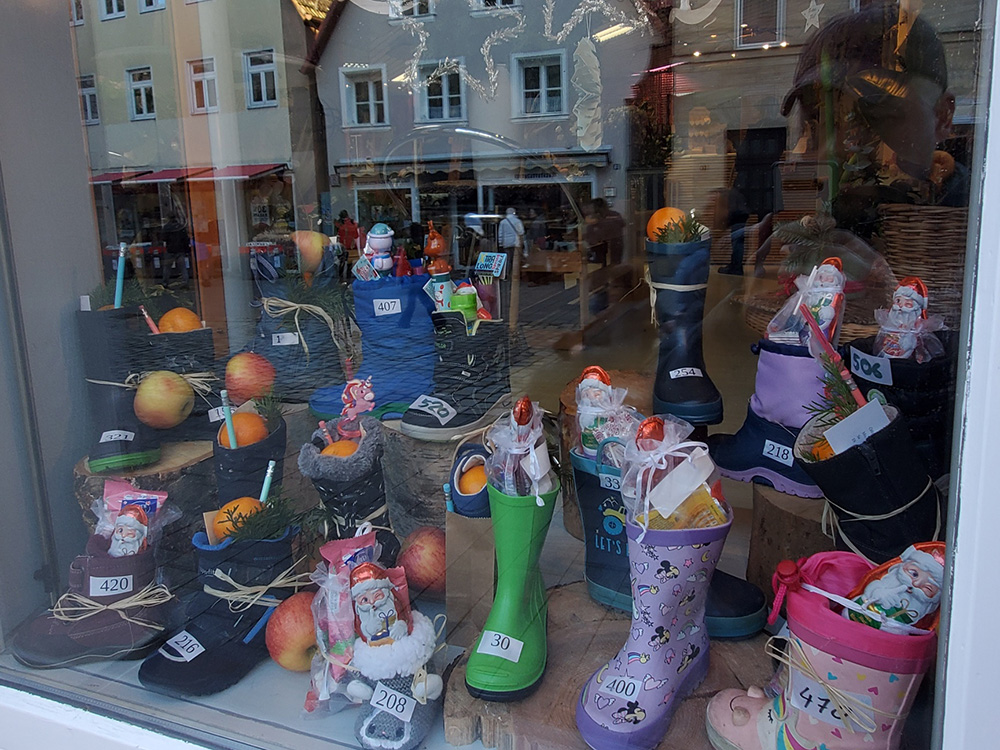 Im Schaufenster sind verschiedene bunte Weihnachtsstiefel aus Gummi und Stoff aufgestellt, die mit kleinen Geschenken, Süßigkeiten und Orangen gefüllt sind. Die Stiefel haben unterschiedliche Farben und Muster, darunter grün, lila mit Sternen, schwarz und rosa. Einige Stiefel tragen kleine Zettel mit Nummern. Im Hintergrund spiegeln sich Häuser und Passanten auf der Straße.