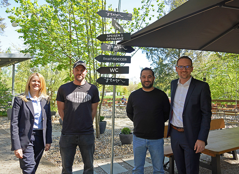 Im Vordergrund steht eine Wegweisertafel mit mehreren rechteckigen Schildern, die in verschiedene Richtungen zeigen. Die Schilder tragen weiße Schrift auf dunklem Grund und weisen unter anderem auf 'Speisen & Getränke', 'Beach Soccer' und 'WC' hin. Im Hintergrund sind Bäume mit frischem Laub und mehrere Holzbänke und -tische zu sehen. Vier Personen stehen um die Tafel, zwei davon tragen Anzüge, eine Person trägt ein schwarzes T-Shirt und eine weitere eine dunkle Jacke. Die Szene spielt im Freien bei Tageslicht, ein Teil des Bereichs ist durch ein Sonnensegel beschattet.