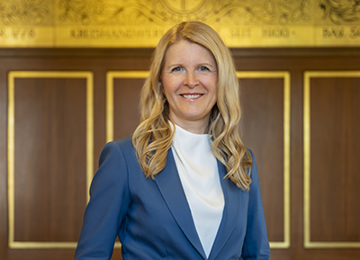 Frau mit langen, gewellten blonden Haaren trägt weißen Rollkragenpullover und blauen Blazer vor einer Holzwand mit goldfarbenem Dekor