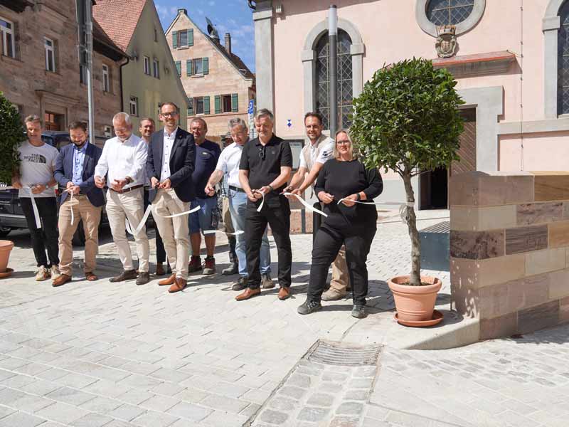 Eine Gruppe von Menschen steht auf einem Platz und schneidet gemeinsam ein Band durch. Dieses ist von einem Baum in einem Kübel zu einem anderen aufgespannt. Rechts sieht man einen teil einer Mauer, hinter der Gruppe einen rosafarbenen Kirchenbau.