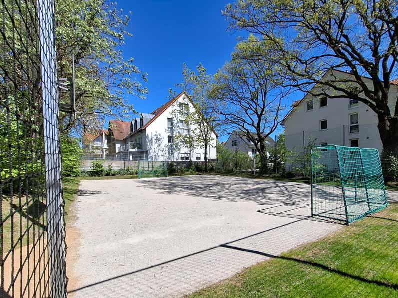 Ein Bolzplatz mit zwei Toren und einem Zaun darum. Im Hintergrund sieht man Wohnhäuser und große Bäume