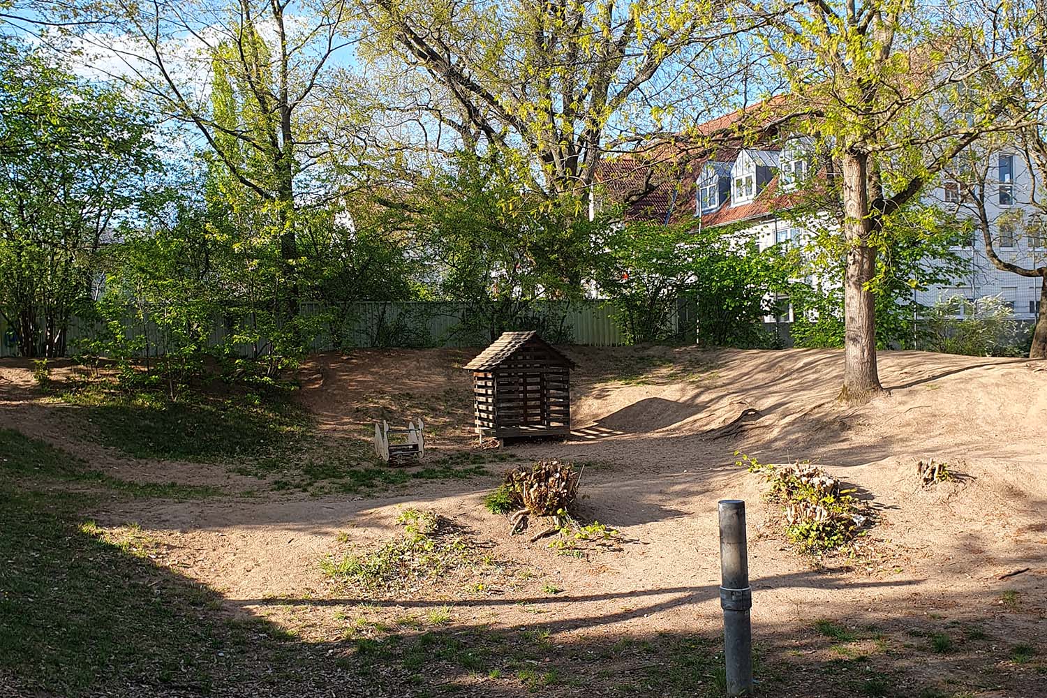 Eine Freifläche mit sandigem Boden und vereinzelten Sträuchern. In der Mitte steht ein kleines hölzernes Spielhaus. Dahinter befinden sich Bäume mit frischem Laub sowie Wohngebäude im Hintergrund.