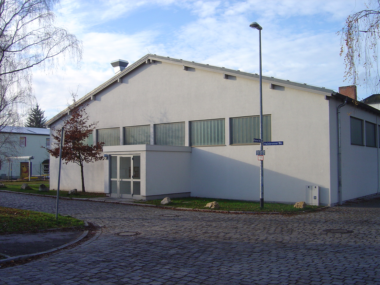 Ein einstöckiges, weiß gestrichenes Gebäude mit flachem Satteldach steht an einer gepflasterten Straßenecke. Die Fassade ist schlicht, mit mehreren länglichen Fenstern im oberen Wandbereich. Vor dem Eingang befindet sich ein kleiner, vorgebauter Windfang mit Glastüren. Ein Laternenmast und ein Straßenschild stehen am Gehweg. Im Vordergrund sind kahle Bäume und ein paar große Steine am Straßenrand zu sehen. Der Himmel ist leicht bewölkt.
