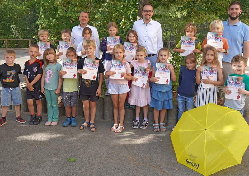 Eine Gruppe von Kindern steht auf einem Hof und alle halten Urkunden in den Händen. Hinter ihnen stehen drei Männer. Und vor ihnen liegt ein aufgespannter gelber Regenschirm.