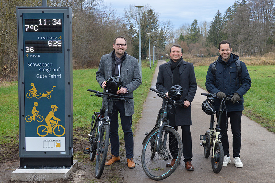 Eine dunkelblaue Säule zeigt Zahlen an, unter anderem die aktuelle Temperatur, die Uhrzeit und die Anzahl der erfassten Radfahrer. Daneben stehen drei Männer neben ihren Fahrrädern.